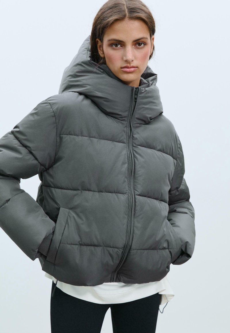 Puffer jacket in dark gray, featuring a high collar, horizontal quilted sections, a front zipper, and side pockets. Textured fabric for warmth.