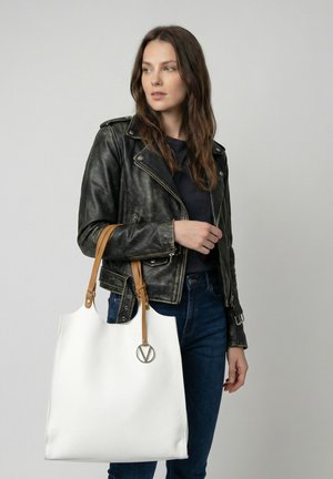 Kvinde med langt brunt hår iført sort læderjakke og jeans, der holder en stor hvid mulepose med beige håndtag og metal logo charm.