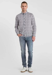 Chemise blanche à boutons avec un motif abstrait noir, combinée avec un jean bleu clair et des baskets blanches, présentée sur un modèle masculin.