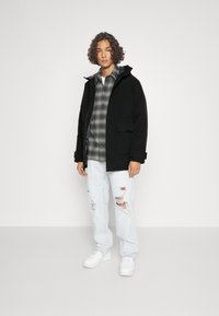 Giacca nera con cappuccio, chiusura con zip, due tasche frontali. Sotto, una camicia a quadri grigia. Indossata con jeans azzurri chiari strappati e sneakers bianche.