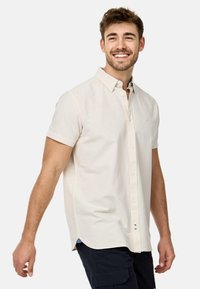 Camisa de botones de manga corta en un ligero tono beige con cuello de punta y un sutil logotipo. Presenta un dobladillo recto y una textura de tela suave.