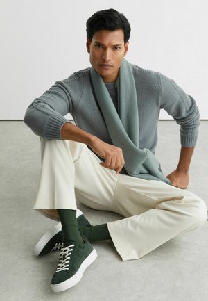 Hellgrauer Strickpullover mit strukturierten Ärmeln, mintgrüner Schal, cremefarbene Hose und dunkelgrüne Wildledersneaker mit weißen Schnürsenkeln.