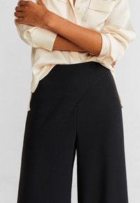 Sorte bredde ben bukser lavet af glat stof med et asymmetrisk front sømdetalje. Parret med en lysfarvet, løstsiddende blouse.