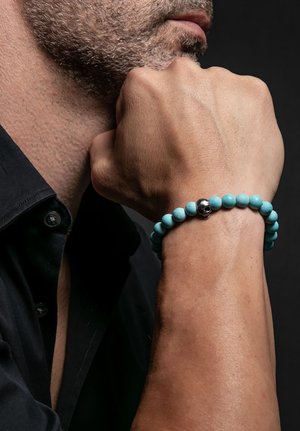 Bracelet en perles turquoise avec un accent en argent en forme de skull. Texture lisse, forme ronde, porté au poignet à côté d'une main reposant sur un menton.