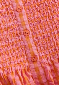 Roza in oranžna smockana tkanina s horizontalnim nabiranjem, ki vsebuje dve oranžni gumb in kariraste teksturne poudarke.