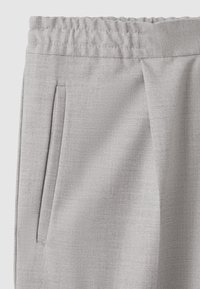 Ceinture de pantalon gris clair avec élastique froncé, une poche latérale verticale et une texture subtile du tissu visible sur un fond uni.