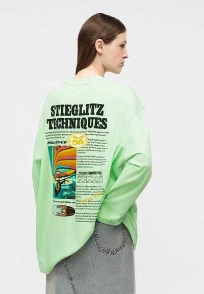 Γυναίκα με μακριά καστανά μαλλιά που φοράει ανοιχτό πράσινο φούτερ με πολύχρωμα γραφικά και κείμενο στο πίσω μέρος, συνδυασμένο με φούστα από τζιν.