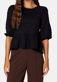 Haut texturé noir avec manches trois-quarts bouffantes, taille froncée et design peplum, assorti à un pantalon marron ajusté.