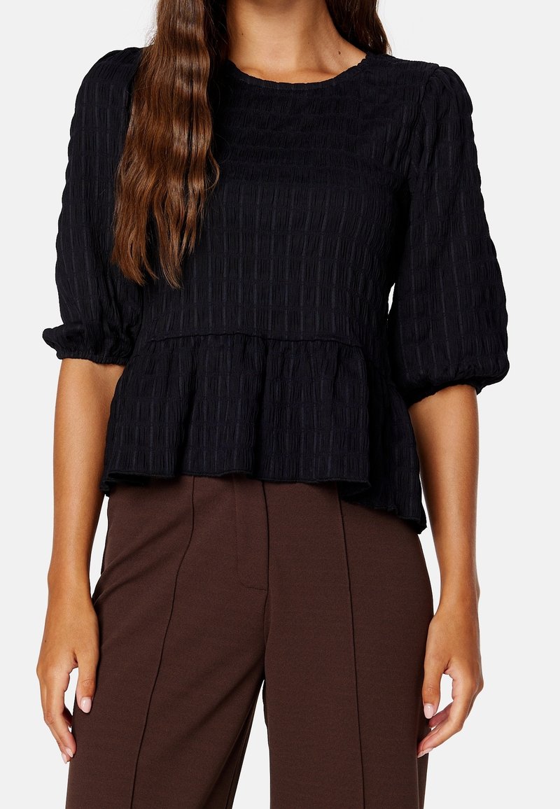 Haut texturé noir avec manches trois-quarts bouffantes, taille froncée et design peplum, assorti à un pantalon marron ajusté.