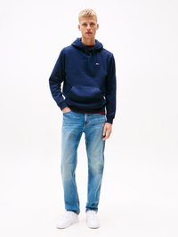 Young man wearing a navy hoodie, light blue jeans, and white sneakers standing against a plain white background.