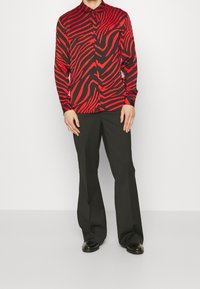 Maglia a maniche lunghe rossa e nera con motivo a strisce zebrato, colletto abbottonato e texture liscia, abbinata a pantaloni e scarpe nere.