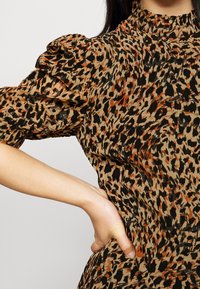 Ruskea ja musta leopardikuosisena blusa puffi varrukatega, kõrge kaelusega ning kogutud tekstuuriga. Tuntavad detailid hõlmavad oranži aktsenti.