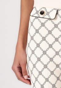 Kremna krilo z črnim geometrijskim vzorcem, gladka tekstura, stranska žepa z zlatim gumbom kot poudarkom. Poudarjen stik z roko.