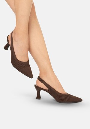 Piernas que llevan zapatos marrones de malla con tacón kitten y tira trasera, con punteras puntiagudas, sobre un fondo gris claro.