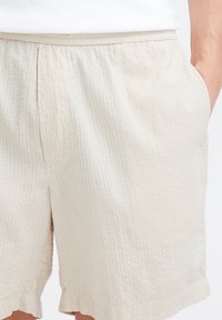 Pantaloni scurți bej texturați, cu talie elastică, buzunare laterale și margini nefinisate, având un model subtil cu dungi verticale.