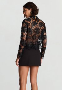 Top in pizzo nero con ricamo floreale, maniche lunghe, orlo smerlato e dettaglio con apertura a goccia sul retro, abbinato a una gonna nera semplice.