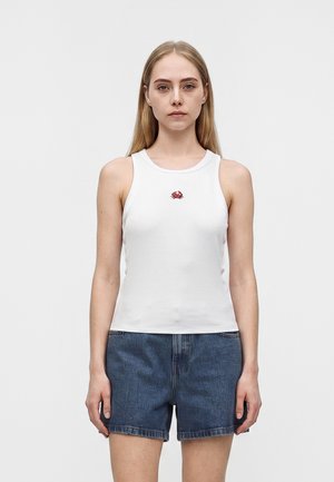 Jeune femme debout face à l'avant, portant un haut blanc sans manches avec un petit emblème de crabe rouge et un short en jean bleu, fond neutre.