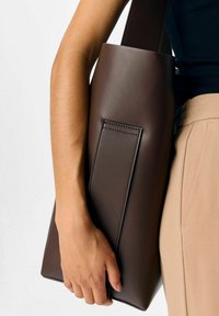 Personne portant un pantalon beige et un haut noir tenant un grand sac fourre-tout en cuir marron foncé par sa anse sur un fond blanc.