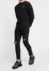 Felpa nera con polsini a coste e logo sul petto, abbinata a jeans neri strappati con dettagli rotti al ginocchio e scarpe da ginnastica nere.