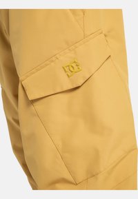 Pantaloni gialli impermeabili con una tasca laterale caratterizzata da un evidente logo DC. Il tessuto ha una consistenza liscia e un design strutturato.