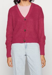 Knitted cardigan in fuchsia with a v-neck, long sleeves, three large brown buttons, and a cropped design. Worn over a light pink top.