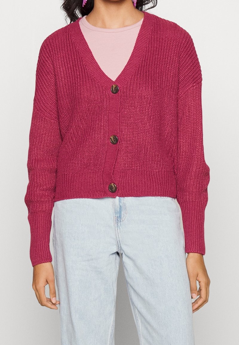 Knitted cardigan in fuchsia with a v-neck, long sleeves, three large brown buttons, and a cropped design. Worn over a light pink top.