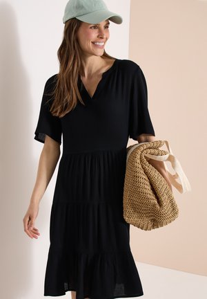 Mujer sonriendo, vistiendo un vestido negro de manga corta y una gorra verde claro, sosteniendo un gran bolso tejido color marrón claro con asas claras.