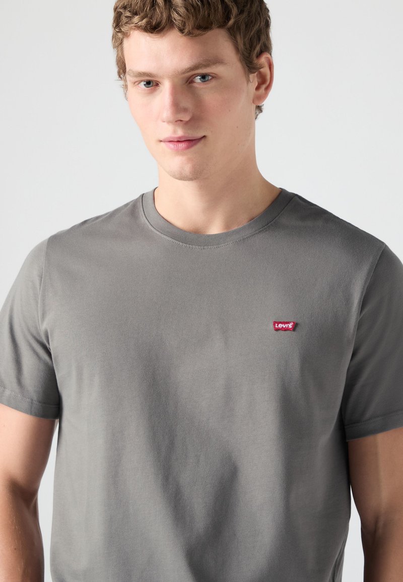 Jeune homme aux cheveux courts et bouclés portant un t-shirt gris uni Levi's, regardant vers l'avant sur un fond neutre.
