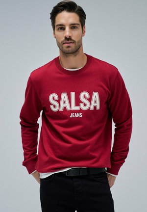 Sudadera roja con el texto en blanco "SALSA JEANS" en contraste. Cuello y puños de canalé, combinada con jeans negros. Diseño simple, corte informal.
