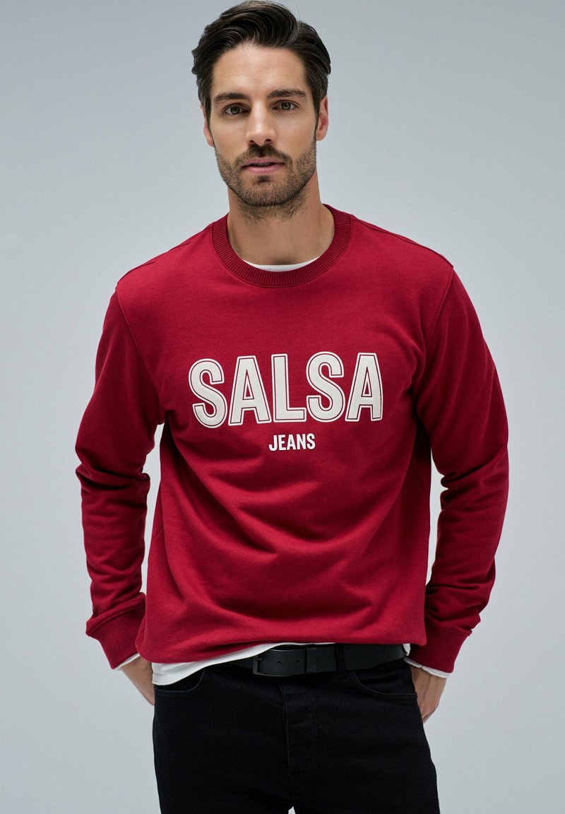 Sweat-shirt rouge avec le texte blanc contrastant "SALSA JEANS". Col et poignets côtelés, accompagné de jeans noirs. Design simple, coupe décontractée.