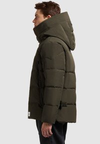 Tamsiai žalia pūkinė striukė su aukštu apykakliu ir kapišonu, su siuvinėta tekstūra, šonine užtrauktuku kišenėje ir reguliuojama juosmens detale.