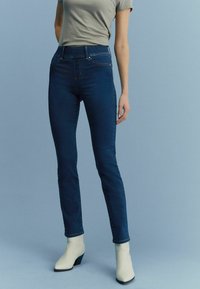 Slim-fit donkerblauwe denim jeans met een hoge taille, subtiele stiksels en achterzakken. Gecombineerd met off-white enkellaarzen.