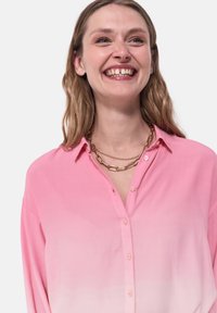 Blusa button-up rosa chiaro con effetto degradé, texture morbida e maniche lunghe. Accentuata con una collana di catena dorata.