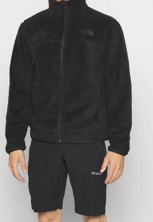 Man wearing a black North Face zip-up fleece jacket and black Columbia shorts standing against a plain background.
