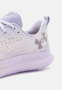 Close-up van een lavendelkleurige Under Armour hardloopschoen met een gebreid bovenmateriaal, witte veters en "FLOW" geperst op de tussenzool.
