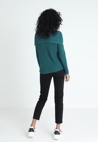 Pull bleu sarcelle à texture côtelée avec un large col, assorti à un pantalon noir moulant et des baskets blanches avec des accents rouges et noirs.