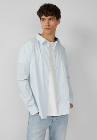 Lichtblauw overhemdd met een subtiele bloemenprint, lange mouwen en een klassieke kraag, gecombineerd met een wit T-shirt en lichte jeans.