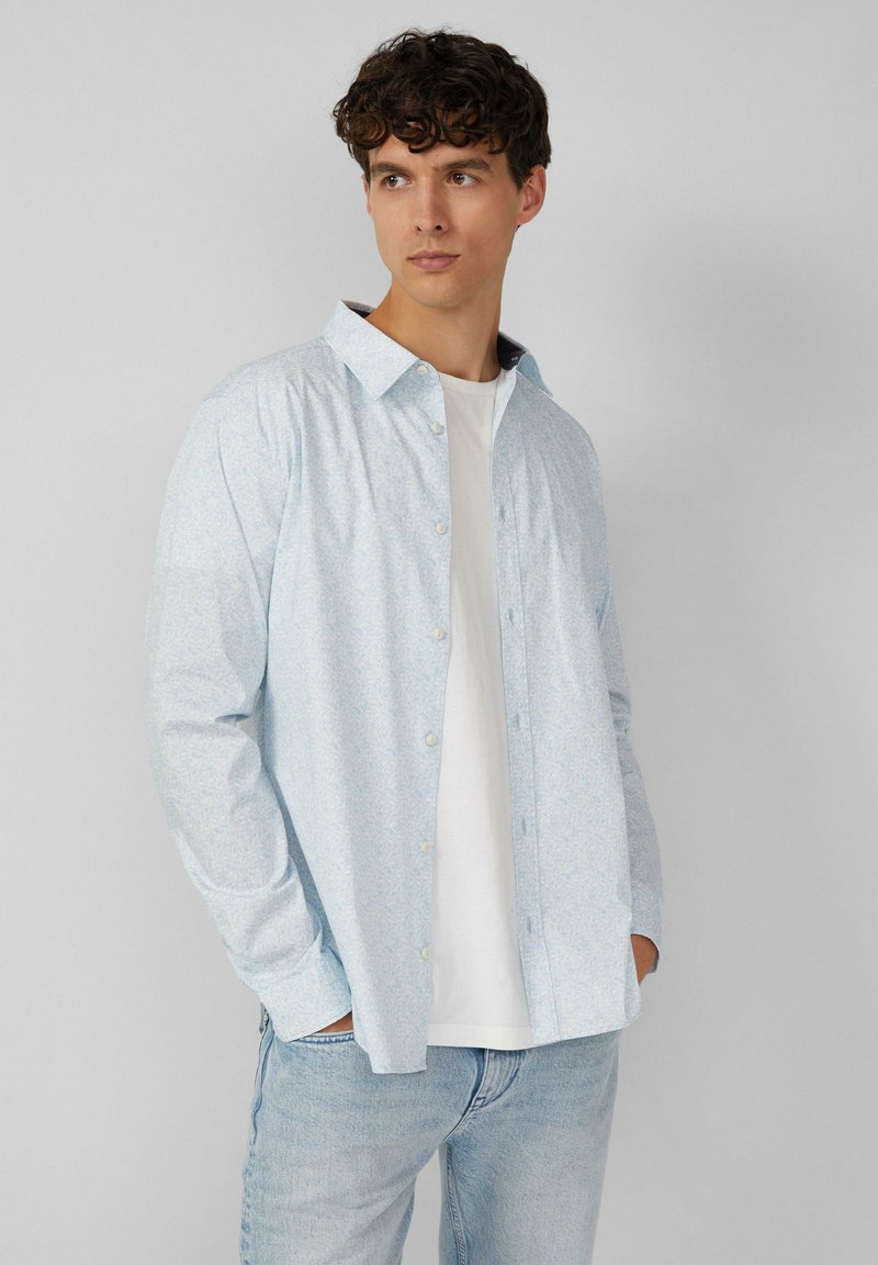 Lichtblauw overhemdd met een subtiele bloemenprint, lange mouwen en een klassieke kraag, gecombineerd met een wit T-shirt en lichte jeans.