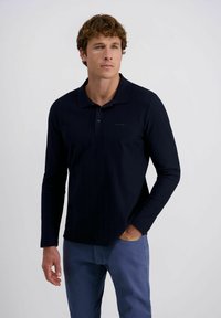 Mann mit lockigem Haar, der ein schwarzes Langarm-Polohemd und blaue Hosen trägt, eine Hand in der Tasche, leicht nach rechts schauend.