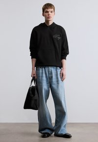 Μαύρο φούτερ με κουκούλα και λογότυπο «Hugo», άνετη εφαρμογή, σε συνδυασμό με φαρδιά τζιν παντελόνια σε ανοιχτό μπλε χρώμα. Το μοντέλο κρατάει μια μαύρη τσάντα ώμου και φοράει μαύρα παπούτσια.