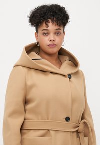 Beige hettejakke med innsvingt midje, to frontlommer og en enkelt svart knapp. Myk tekstur, minimalistisk design.