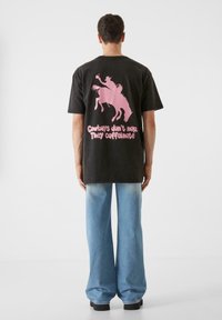 Črna bombažna majica s kratkimi rokavi z roza grafičnim dizajnom, ki prikazuje kavboja na konju in besedilo. Utemeljena z svetlimi jeans hlačami širokega kroja.