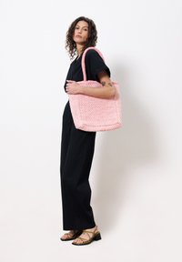 Roze geweven draagtas in vierkante vorm, met dubbele hengsels en een gestructureerd oppervlak. Model houdt de tas vast terwijl ze een zwarte outfit en bandjesandalen draagt.