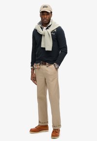 Marineblauwe trui met logo, beige chinos en tan laarzen. Laagjes met een geruit overhemd eronder, lichtgrijze gebreide sjaal en een lichtbeige pet.