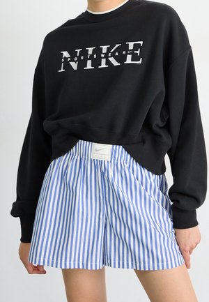 Persoon draagt een zwarte Nike trui met wit logo en blauw-wit verticale gestreepte Nike short met logo op de tailleband.