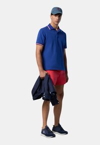 Polo blu con dettagli rossi e bianchi, pantaloni corti rossi, giacca blu navy tenuta in mano e sneakers navy. Cappellino in denim chiaro con logo.