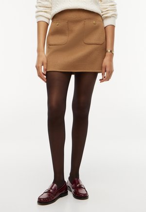 Femme portant une mini-jupe camel avec des poches à l'avant, des collants noirs, des mocassins bordeaux, un pull blanc et une montre-bracelet en or.