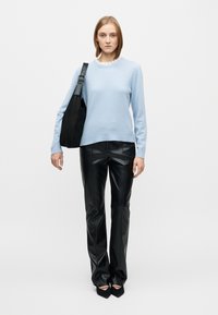 Pull bleu clair à col côtelé, associé  à un pantalon en cuir noir et un sac à bandoulière noir ; le mannequin pose devant un fond blanc uni.