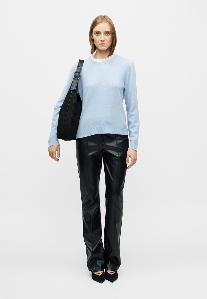 Pull bleu clair à col côtelé, associé à un pantalon en cuir noir et un sac à bandoulière noir ; le mannequin pose devant un fond blanc uni.