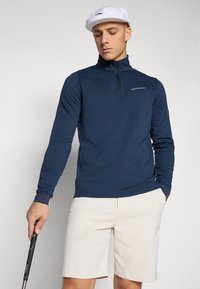 Marineblauwe longsleeve pullover met halve ritssluiting en aansluitende manchetten, gecombineerd met lichtbeige shorts. Glad materiaal met logo op de borst.